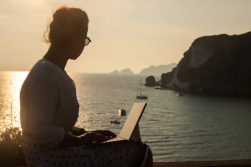 Frau mit Laptop arbeitet. Im Hintergrund eine traumhafte Kulisse am Meer. Ihre Frage: Ich habe keine Erfahrung auf dem Wasser. Kann ich trotzdem teilnehmen?